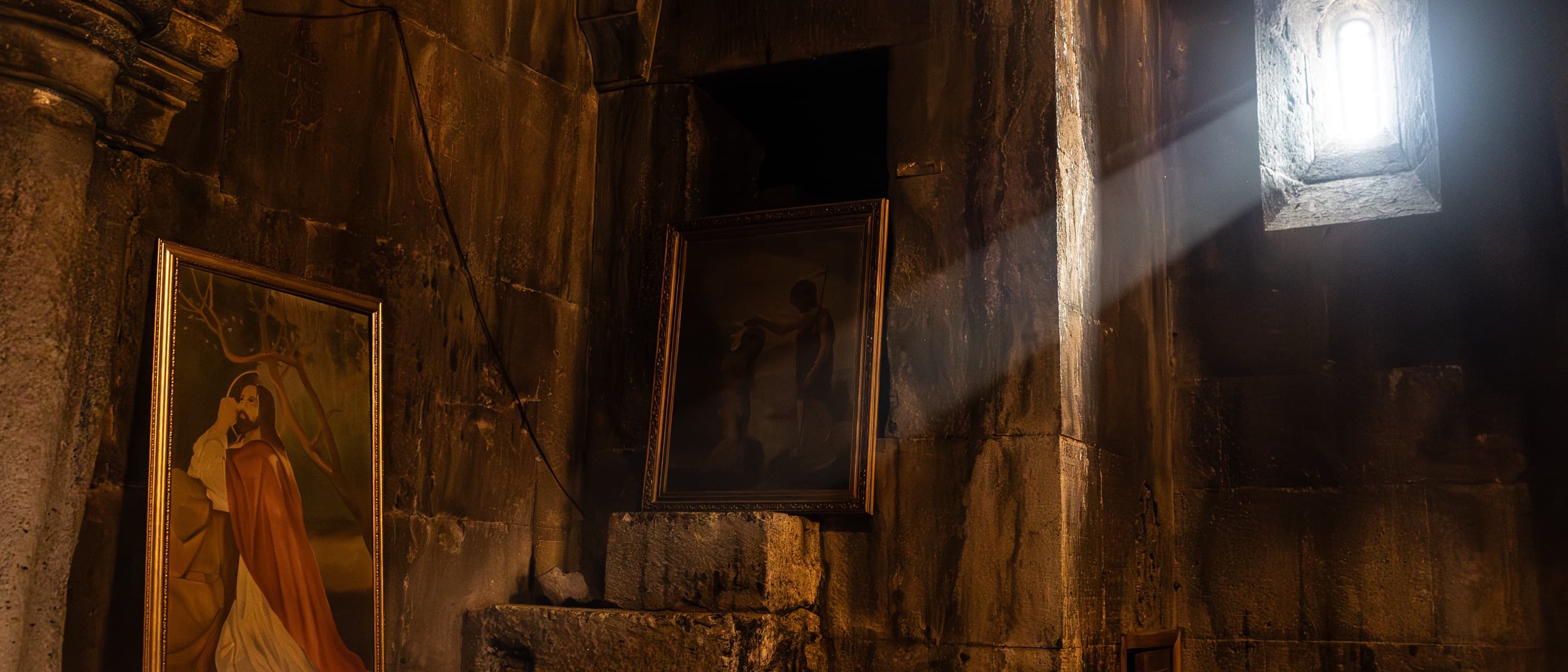 Un haz de luz entra por el ventanuco de un monasterio armenio y cruza la pared de piedra, con dos iconos apoyados abajo.