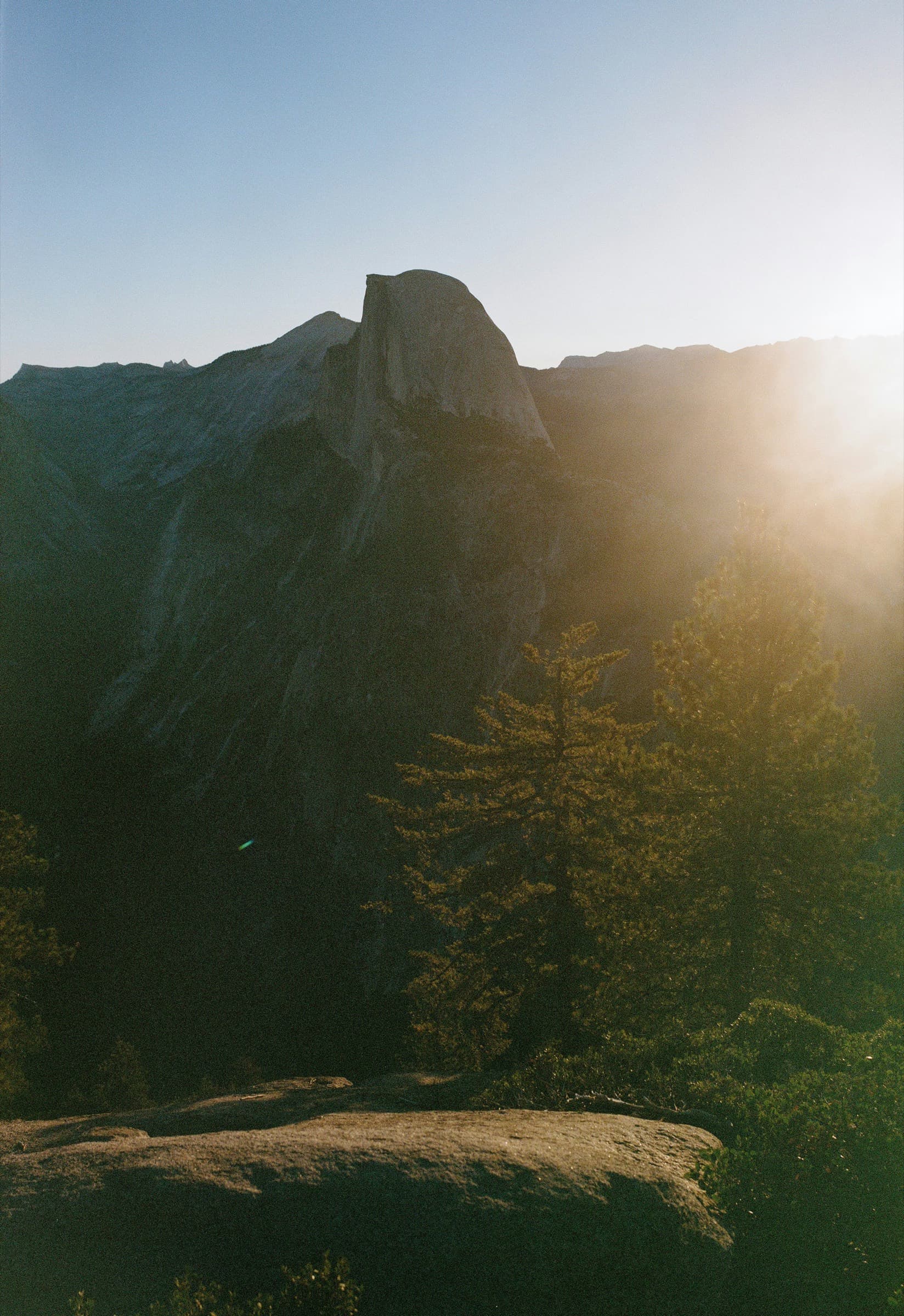 Half Dome de perfil desde Glacier Point al amanecer, con el sol saliendo justo detrás y los pinos a contraluz.