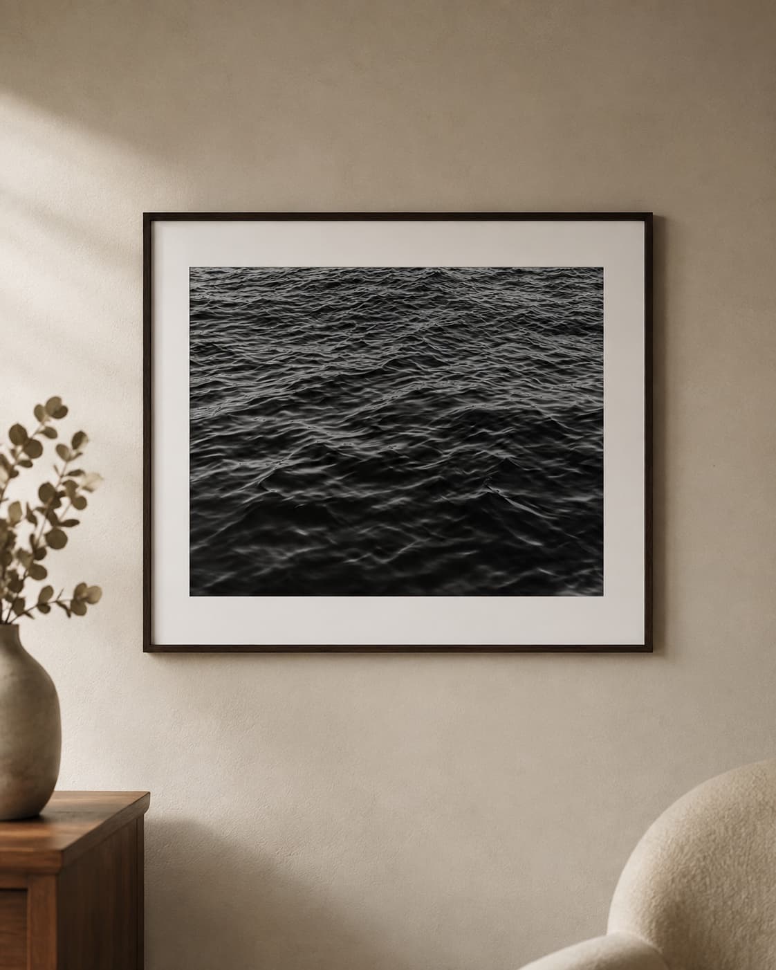 Una impresión enmarcada de agua oscura colgada en una pared cálida, junto a una consola de madera y una rama de eucalipto.